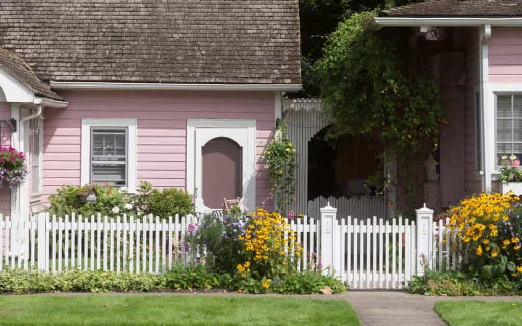 A white stained picket fence accented by flowers and greenery lines the front yard of a pink house